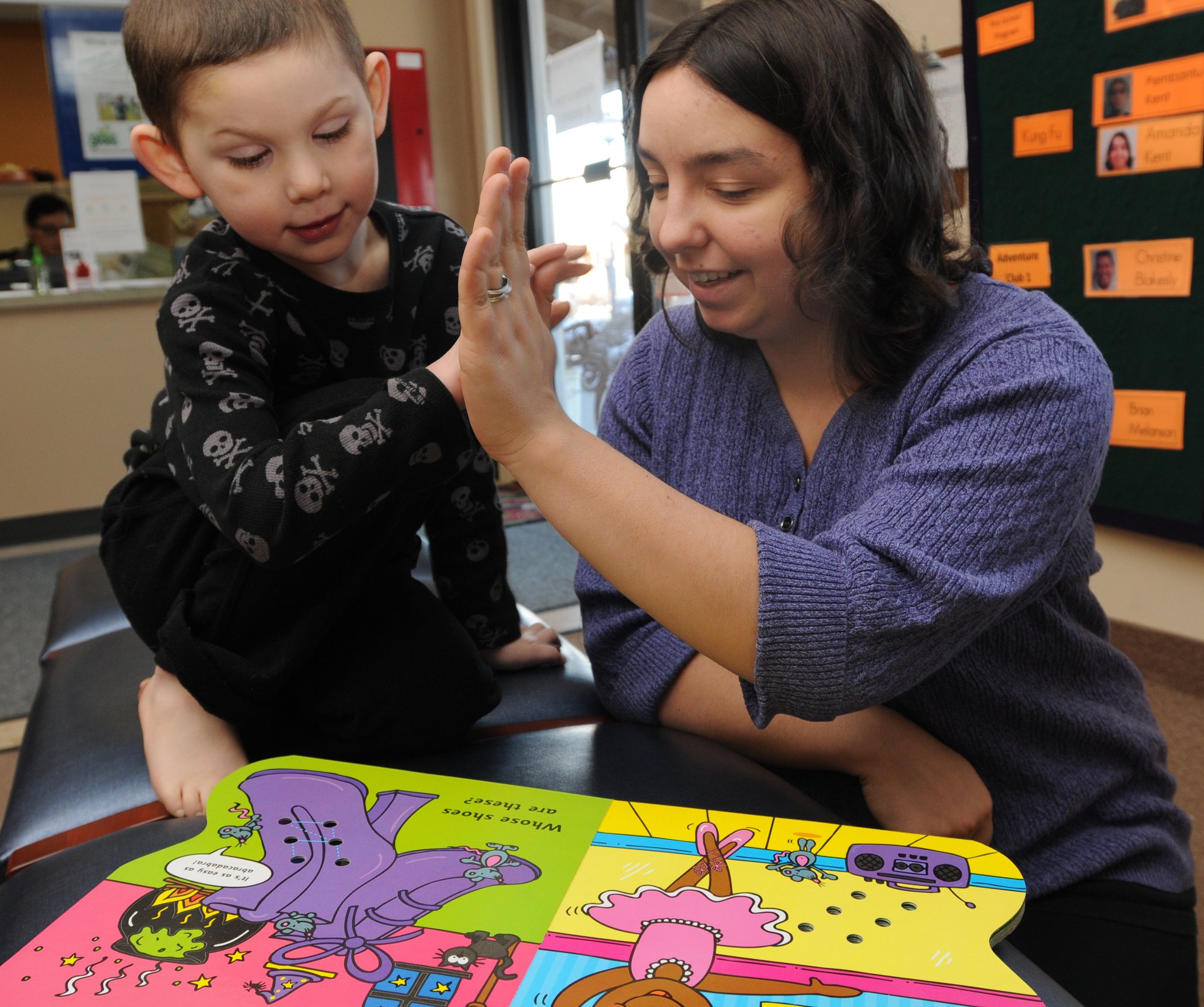 A boy with a disability and a light skin tone high-fives a woman with a light skin tone as they read a picture book together.