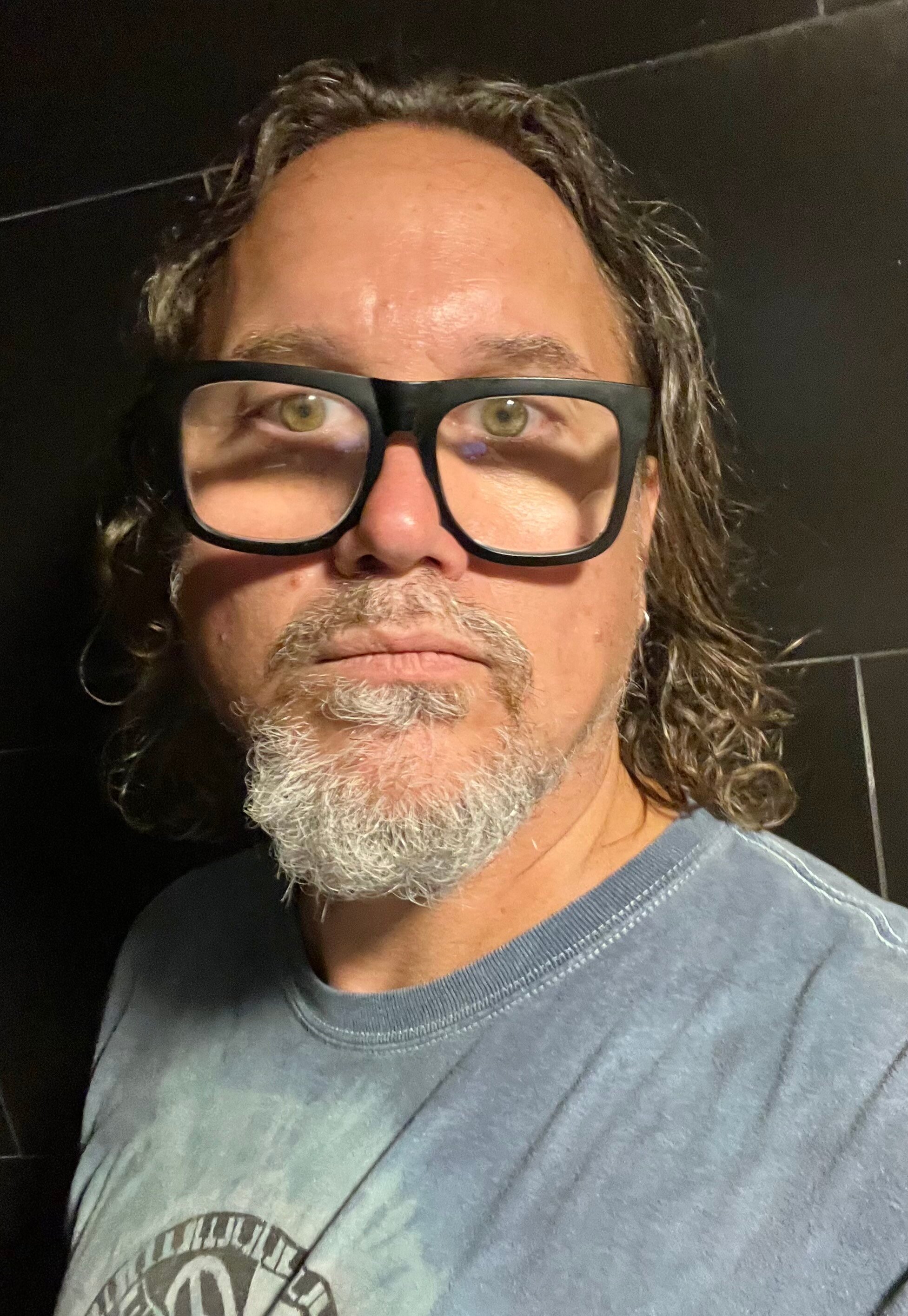 Portrait of Eddy Hougen, who has a light skin tone, a grey goatee and curly brown and grey chin-length hair with glasses.