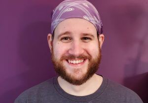 Portrait of Evan Sabourin, who has a light skin tone, a red beard and is wearing a purple bandana while smiling.