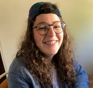 Smiling portrait of Jenna Perna-Elias, who has a light skin tone, curly shoulder-length brown hair, and large glasses.